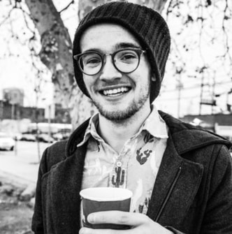 A black and white photo of Quintin, a white male with a slim build, large glasses and a big smile seen from the chest up. He's wearing a beanie and holding a cup.