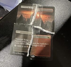 A borderless copy of Haunted Ridge from Innistrad: Midnight Hunt. This card is another victim of Flip It, torn into quarters.